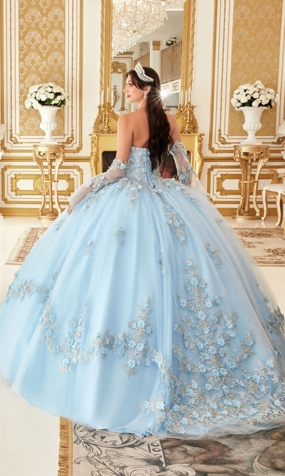 A woman wears the Quinceanera Dress 15714 by Ladivine—a pastel ball gown with silver floral details—standing in an ornate room with gold accents, white flower vases, and a chandelier.